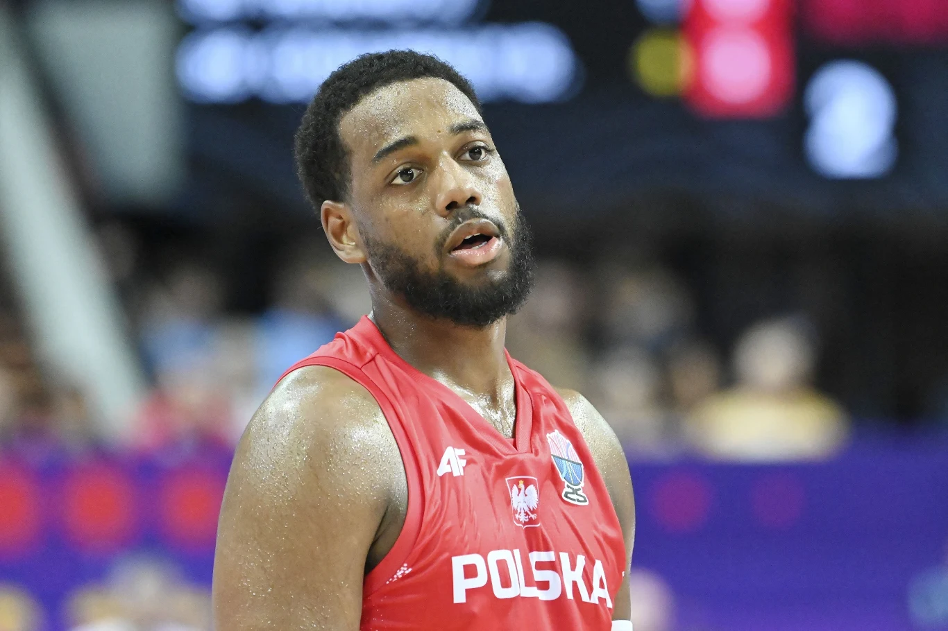 Jordan Loyd Zawodnik polskiej reprezentacji koszykówki w czerwonym stroju sportowym stoi na boisku, w tle rozmyta publiczność i fragment hali sportowej.