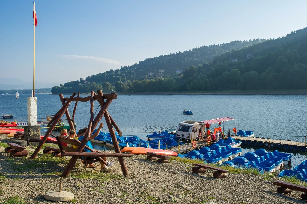 Jezioro Żywieckie to popularne miejsce wypoczynku Plaża przy jeziorze z drewnianą huśtawką na pierwszym planie, licznymi niebieskimi rowerami wodnymi przy pomoście oraz ratownikami na czerwonej łodzi, w tle zalesione wzgórze i zabudowania.