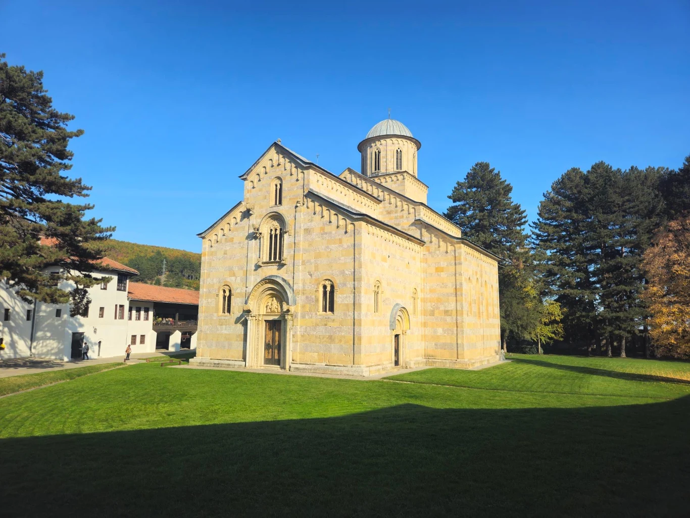 Monaster Wysokie Deczany Kamienny kościół o charakterystycznej architekturze romańskiej otoczony zielonym trawnikiem, pośród wysokich drzew, w słoneczny dzień.