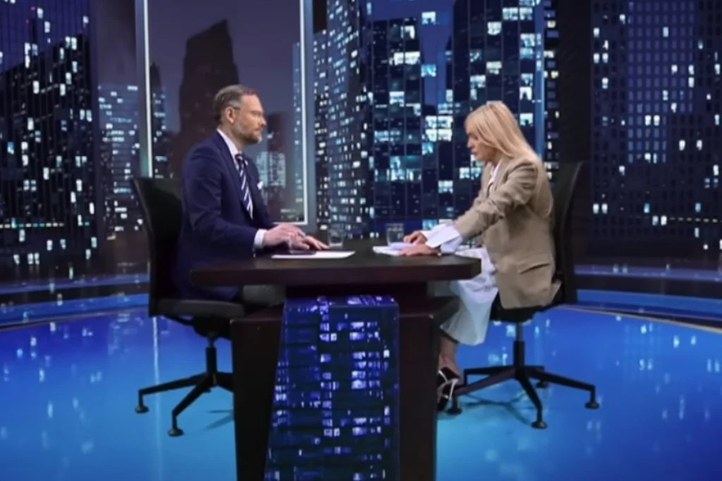 Spięcie Moniki Olejnik z Zbigniewem Boguckim. Poszło o reparacje wojenne od Niemiec Dwójka ludzi siedzi naprzeciw siebie przy stole w studiu telewizyjnym, wokół widoczne są rozświetlone nocą panoramy miasta przez duże okna, mężczyzna ubrany w garnitur, kobieta w jasnej marynarce i białej sukience, oboje prowadzą rozmowę lub wywiad.
