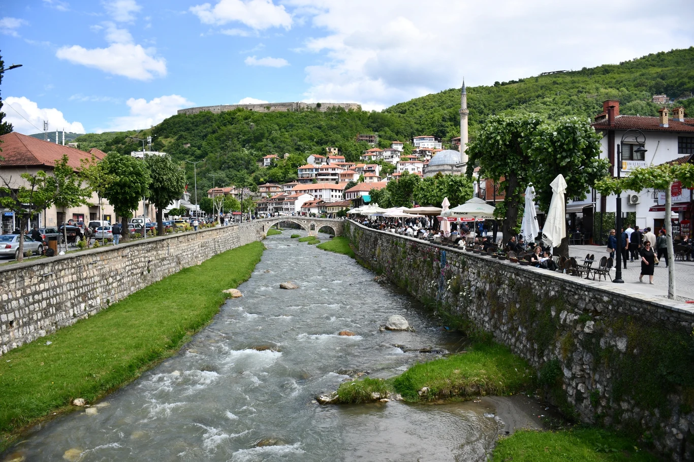 Miasto Prizren Rzeka płynąca przez środek miasta z kamiennym mostem, po obu stronach nabrzeżne uliczki z kawiarniami i ludźmi, w tle zielone wzgórza porośnięte lasem oraz zabudowania miejskie z charakterystycznymi czerwonymi dachami