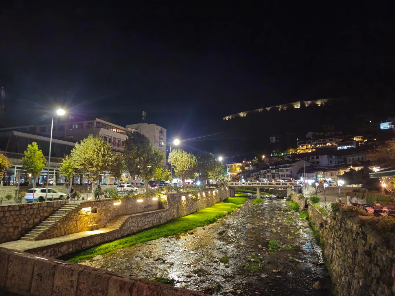 Prizren wieczorową porą Nocny widok na oświetlone centrum miasta z rzeką o niskim stanie wody, otoczoną kamiennymi murkami i drzewami, po lewej stronie nowoczesne budynki oraz szereg zaparkowanych samochodów, na wzgórzu w tle widać iluminowane fragmenty miasta.