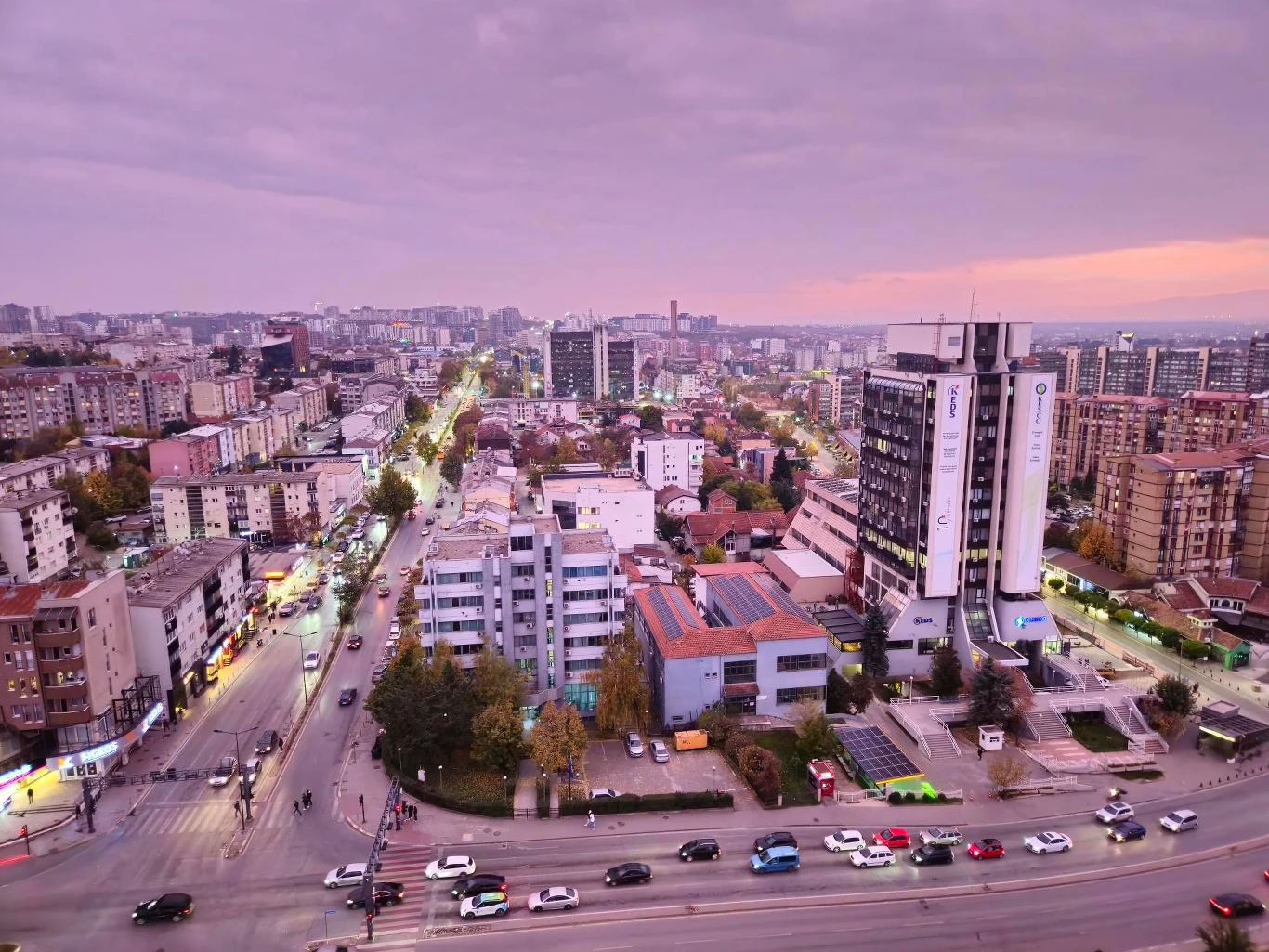 Prisztina Rozległa panorama miasta z licznymi budynkami mieszkalnymi i biurowymi, ruchliwa ulica z samochodami, w tle wieżowce oraz niebo w odcieniach fioletu i pomarańczy podczas zachodu słońca.