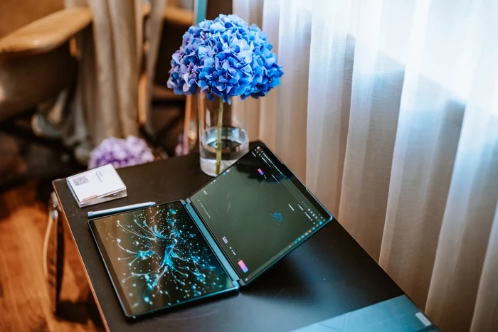 Lenovo Polska Nowoczesny rozkładany laptop na czarnym stoliku obok wazonu z niebieskimi hortensjami i notesu, całość oświetlona naturalnym światłem przez białą firanę