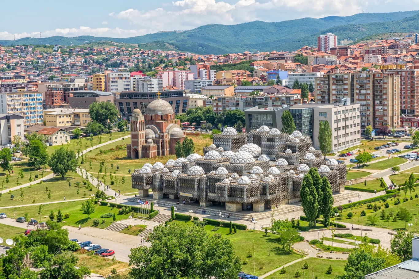 Widok na Bibliotekę Narodową Kosowa i Sobór Chrystusa Zbawiciela w Prisztinie Nowoczesny kompleks budynków o unikalnej architekturze z licznymi kopułami otoczony zielenią i alejkami, w tle panorama miasta z wieloma blokami mieszkalnymi i górzystym krajobrazem.