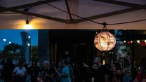 Papierowy lampion i ciepłe światło lampki pod daszkiem na pierwszym planie, w tle grupa ludzi uczestniczących w wieczornym wydarzeniu plenerowym.