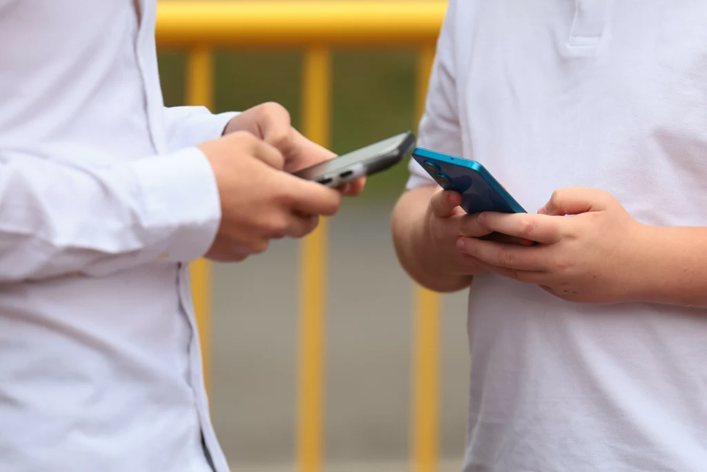 Dwie osoby w białych koszulach korzystają ze smartfonów trzymanych w dłoniach, stojąc naprzeciwko siebie przy żółtej barierce.