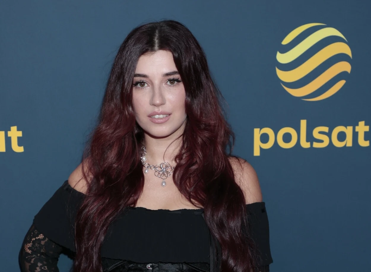 Kobieta o długich, ciemnych włosach w czarnej sukience na tle ściany z logotypem Polsat, ozdobiona delikatną biżuterią na szyi.