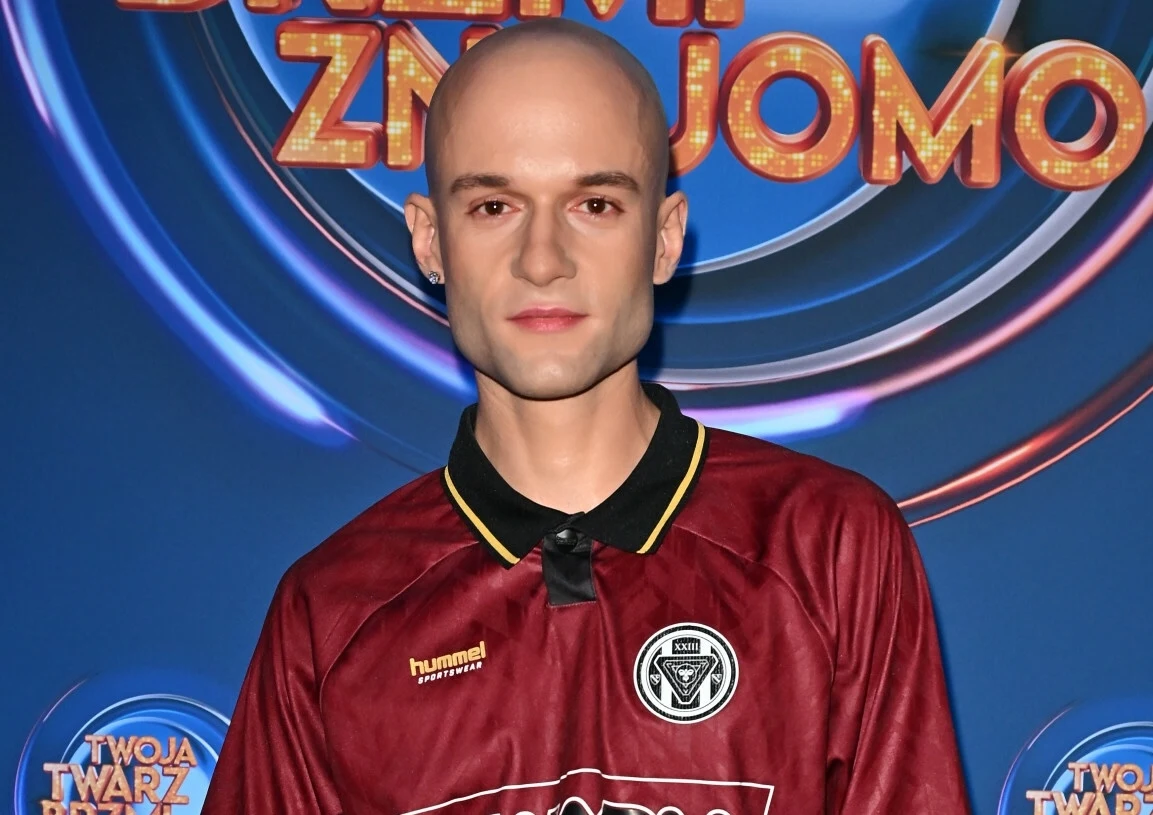Mężczyzna o ogolonej głowie ubrany w bordową koszulkę sportową z logo klubu piłkarskiego, stojący na tle niebieskiej ścianki z kolorowymi napisami.