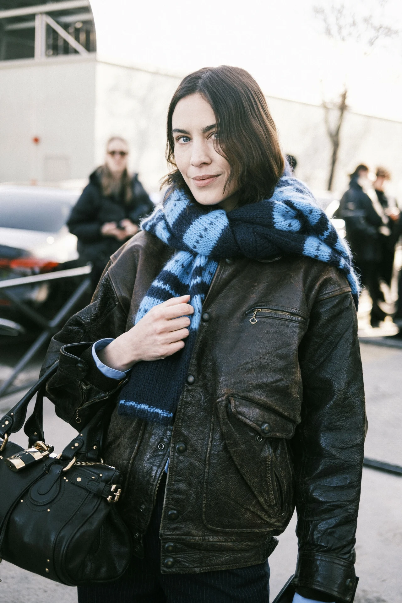 Kobieta o ciemnych włosach, ubrana w skórzaną kurtkę i niebiesko-czarny szalik w geometryczne wzory, trzyma czarną torebkę, stoi na zewnątrz w jasnym świetle dziennym; w tle rozmyte sylwetki innych osób oraz samochody