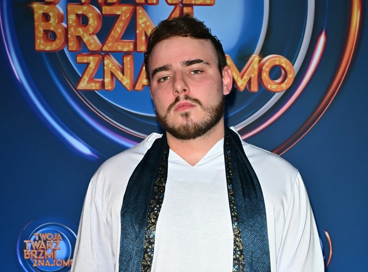 Młody mężczyzna z zarostem ubrany w białą koszulę i granatowy szal stoi na tle ścianki z logo programu 'Twoja twarz brzmi znajomo'.