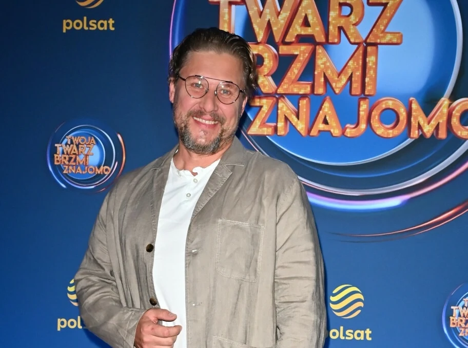 Mężczyzna z brodą i okularami w jasnej marynarce oraz białej koszuli pozuje na tle ścianki promocyjnej programu Twarz Brzmi Znajomo, z widocznym logotypem stacji Polsat.
