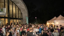 Tłum ludzi zgromadzony przed pawilonem oraz białymi namiotami podczas nocnej imprezy plenerowej, większość uczestników stoi blisko siebie, a w tle widoczne są fragmenty budynku o dużych oknach.