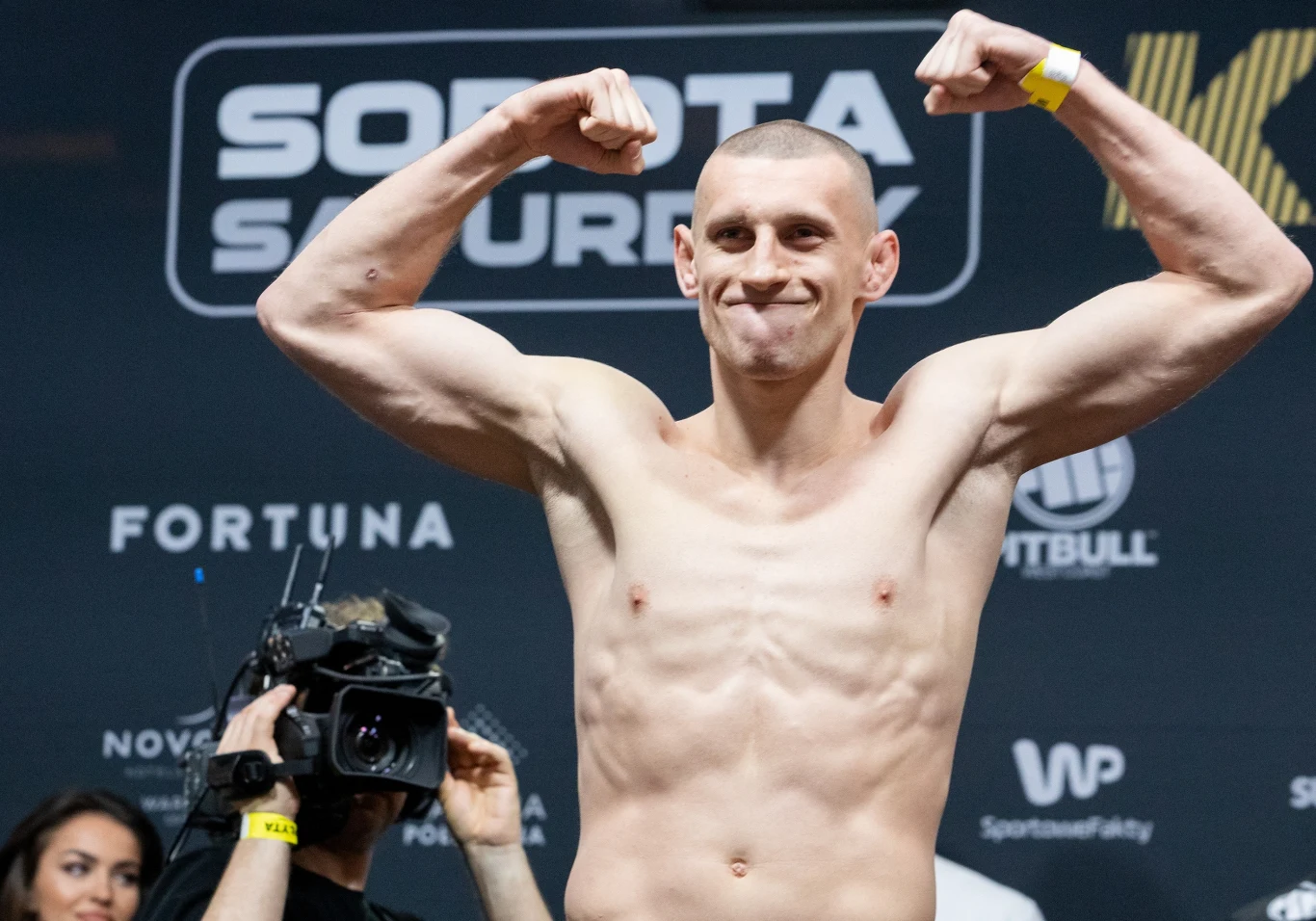 Umięśniony sportowiec bez koszulki prezentuje zaciśnięte pięści i napina ramiona podczas ważenia przed walką, w tle widoczne logo i napisy sponsorów oraz osoby z obsługi wydarzenia.