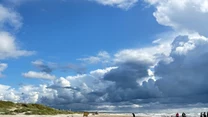 Szeroka, piaszczysta plaża z kilkoma osobami spacerującymi przy brzegu morza, dwa rowery stojące na piasku, dynamiczne chmury na niebie oraz wydmy z zieloną trawą po lewej stronie.