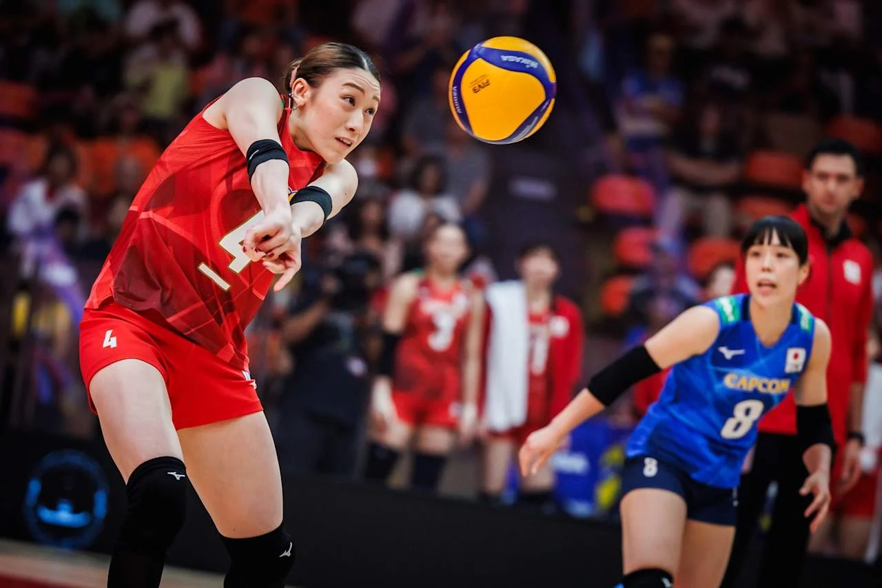 Mayu Ishikawa, kapitan reprezentacji Japonii Siatkarka w czerwonym stroju wykonuje odbicie piłki podczas meczu, w tle druga zawodniczka w niebieskiej koszulce oraz rozmyte sylwetki kibiców na trybunach.