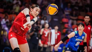 Siatkarka w czerwonym stroju wykonuje odbicie piłki podczas meczu, w tle druga zawodniczka w niebieskiej koszulce oraz rozmyte sylwetki kibiców na trybunach.