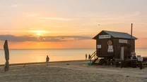 Plaża o zachodzie słońca, na pierwszym planie drewniana budka ratownika z napisem oraz ławki, na piasku widocznych kilka osób spacerujących i odpoczywających nad morzem.