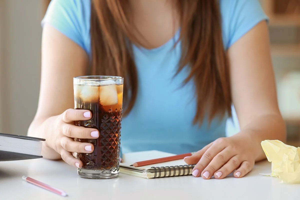 Dłoń kobiety w niebieskiej koszulce trzymająca szklankę z napojem gazowanym i lodem, na biurku leżą otwarty notes, ołówek, książka i zgnieciona kartka papieru.