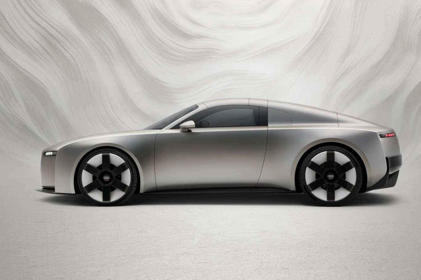 Srebrny samochód sportowy o futurystycznym wyglądzie, z gładkimi liniami karoserii i dużymi, pięcioramiennymi felgami na tle białej, abstrakcyjnej ściany.