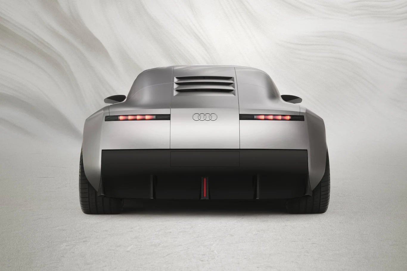 Tylny widok srebrnego samochodu sportowego marki Audi o futurystycznym designie z nowoczesnym oświetleniem i szerokimi kołami na tle minimalistycznego tła.