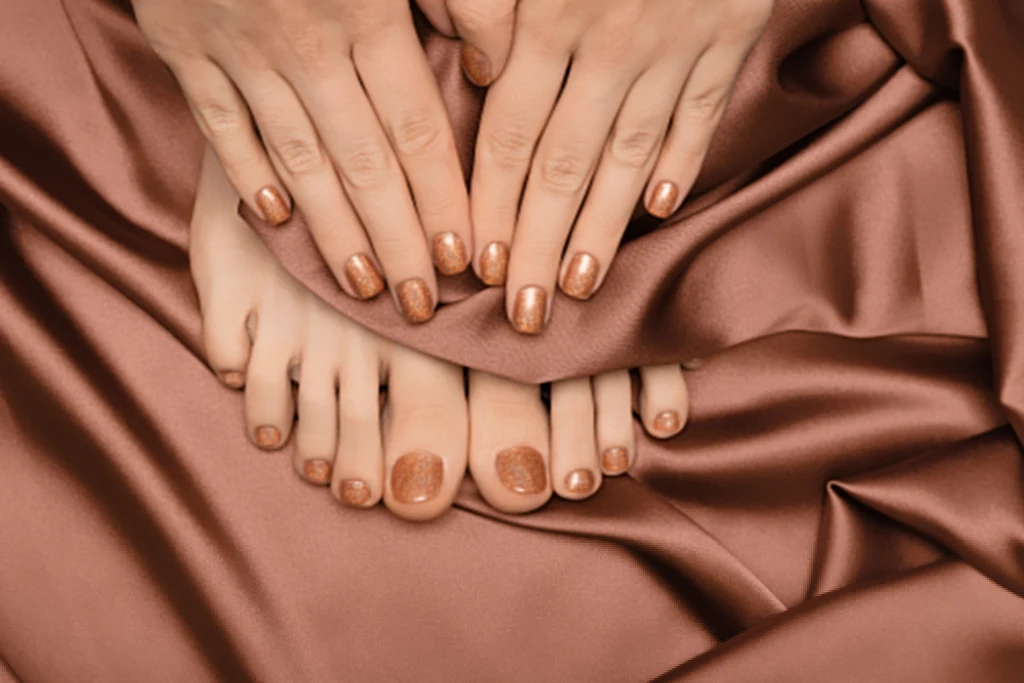 Manicure na jesień 2025 Dłonie i stopy z paznokciami pomalowanymi metalicznym lakierem w tonacji różowo-złotej spoczywają na gładkim, błyszczącym materiale w kolorze dopasowanym do manicure i pedicure.