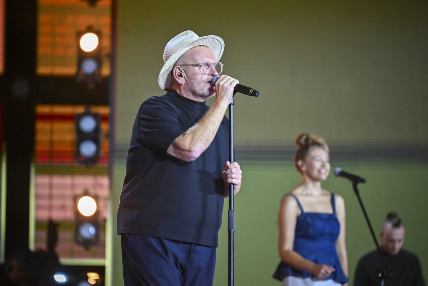 Formacja Nieżywych Schabuff Mężczyzna w okularach i białym kapeluszu śpiewa do mikrofonu na scenie, w tle stoją kobieta oraz inny mężczyzna, widoczne reflektory sceniczne.