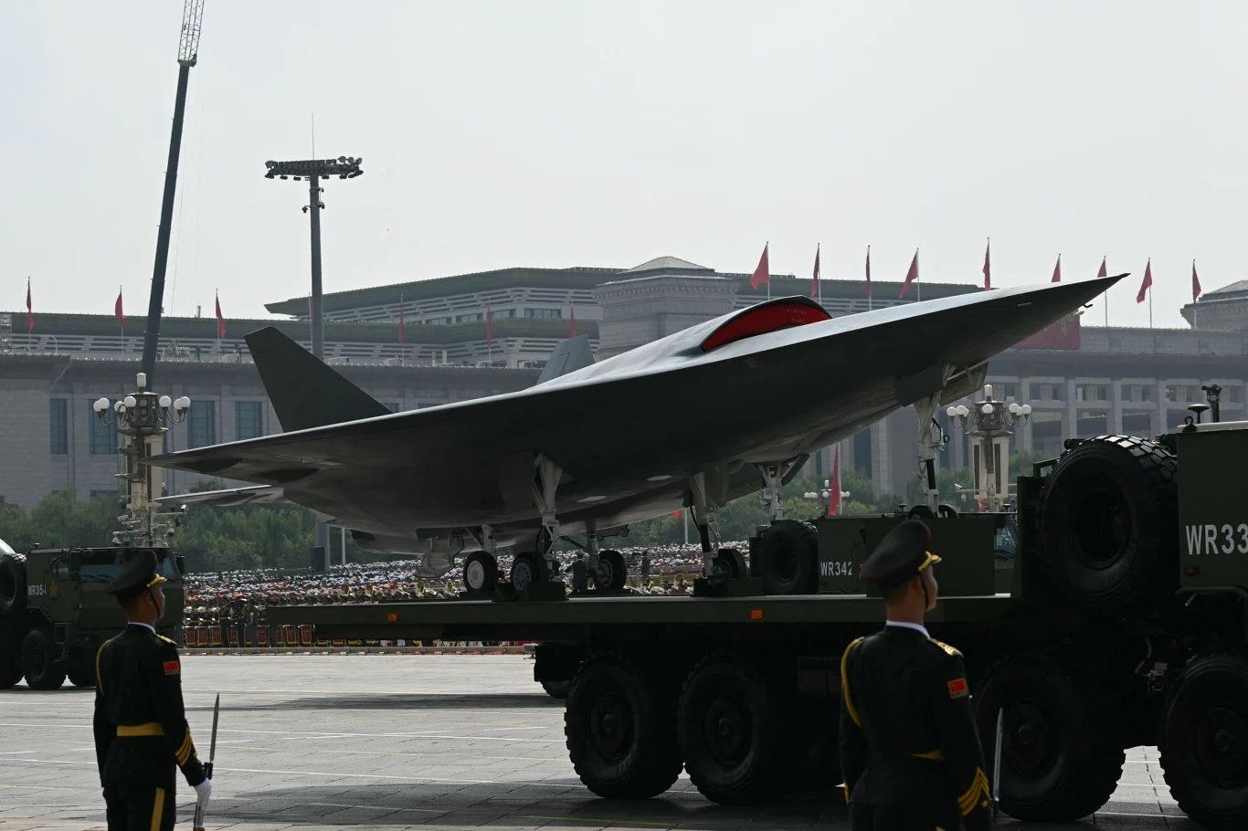 Jeden z zaprezentowanych prototypów CCA Nowoczesny wojskowy dron na platformie transportowej podczas parady, wokół żołnierze w mundurach ceremonialnych, w tle widać trybuny i duży budynek rządowy.