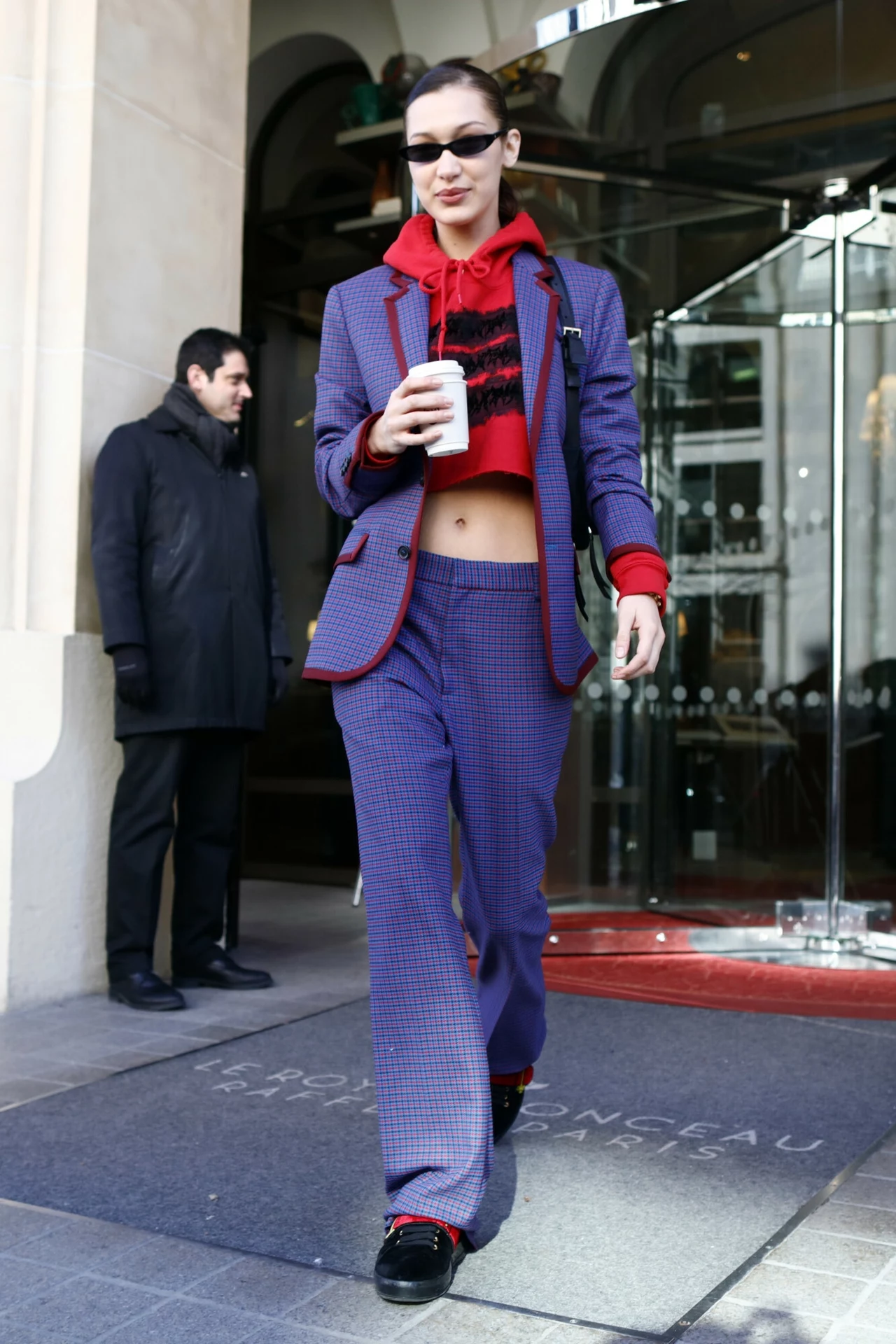 Bella Hadid w fioletowym zestawie, idealnym na jesień Kobieta w modnym garniturze w kratę i czerwonym krótkim swetrze, trzymająca papierowy kubek, wychodzi z hotelu przez szklane drzwi, w tle stoi mężczyzna w czarnym płaszczu.