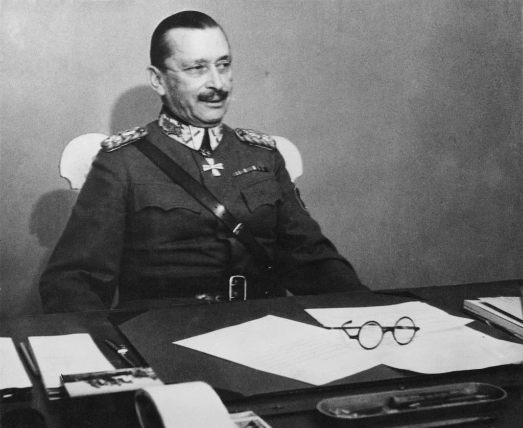 Oficer w mundurze wojskowym z medalami siedzący za biurkiem, na którym leżą okulary, dokumenty oraz przybory do pisania, tło jednolite, brak innych osób.