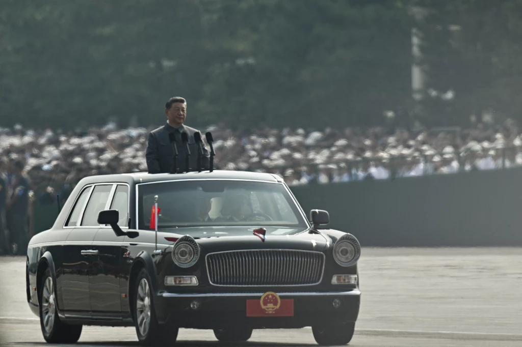 Xi Jiping podczas inspekcji armii na Paradzie Zwycięstwa 3 września 2025 roku Człowiek stojący w otwartym samochodzie biorący udział w oficjalnej paradzie, w tle zgromadzeni ludzie w mundurach oraz widoczne flagi Chin, atmosfera uroczystości państwowej.