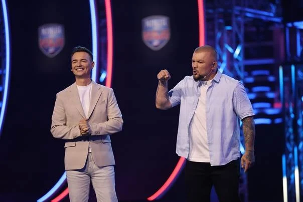 Dwóch mężczyzn na tle widowiskowej scenografii z neonowymi okręgami, jeden ubrany w jasny garnitur, drugi w jasną koszulę z podwiniętymi rękawami energicznie gestykuluje, obaj stoją obok siebie i skupiają uwagę na wydarzeniu na scenie.