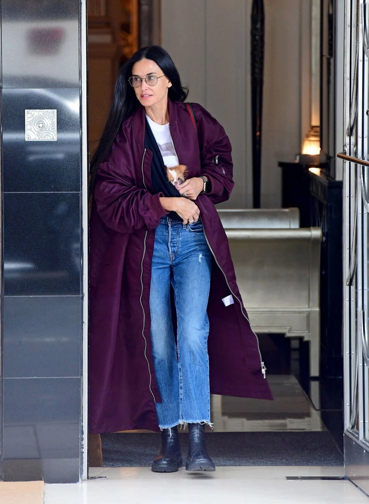 Demi Moore w burgundowym płaszczu Kobieta o długich ciemnych włosach wychodzi przez drzwi w budynku, ubrana w długi bordowy płaszcz, białą koszulkę, niebieskie dżinsy i czarne buty, na twarzy ma okulary.