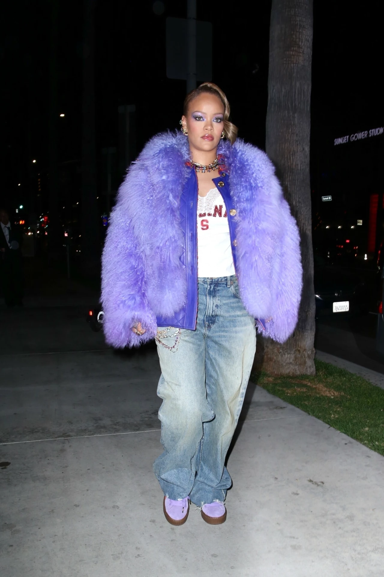 Rihanna w fioletowym futerku Kobieta ubrana w obszerną futrzaną fioletową kurtkę, białą koszulkę oraz szerokie niebieskie jeansy spaceruje nocą chodnikiem miasta. Jej makijaż i dodatki podkreślają ekstrawagancki styl.