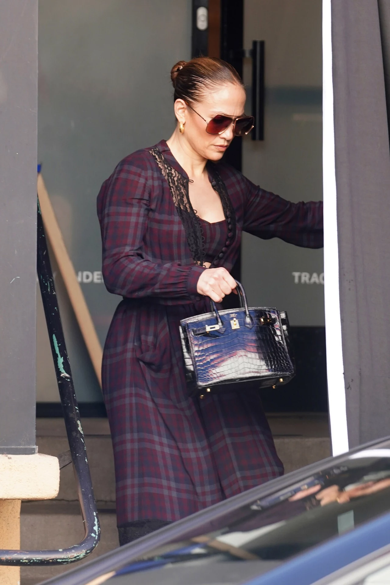 Jennifer Lopez w sukience w kratę Kobieta w ciemnej, kraciastej sukience i okularach przeciwsłonecznych opuszcza budynek, trzymając w ręku luksusową, czarną torebkę.