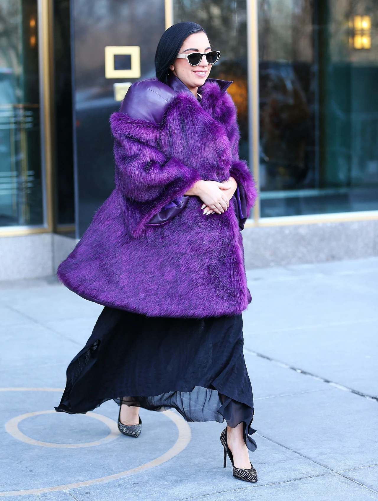 Lady Gaga w fioletowym futrze Kobieta ubrana w fioletowe futro, czarne okulary przeciwsłoneczne oraz czarną, długą sukienkę i szpilki idzie ulicą w miejskiej przestrzeni.