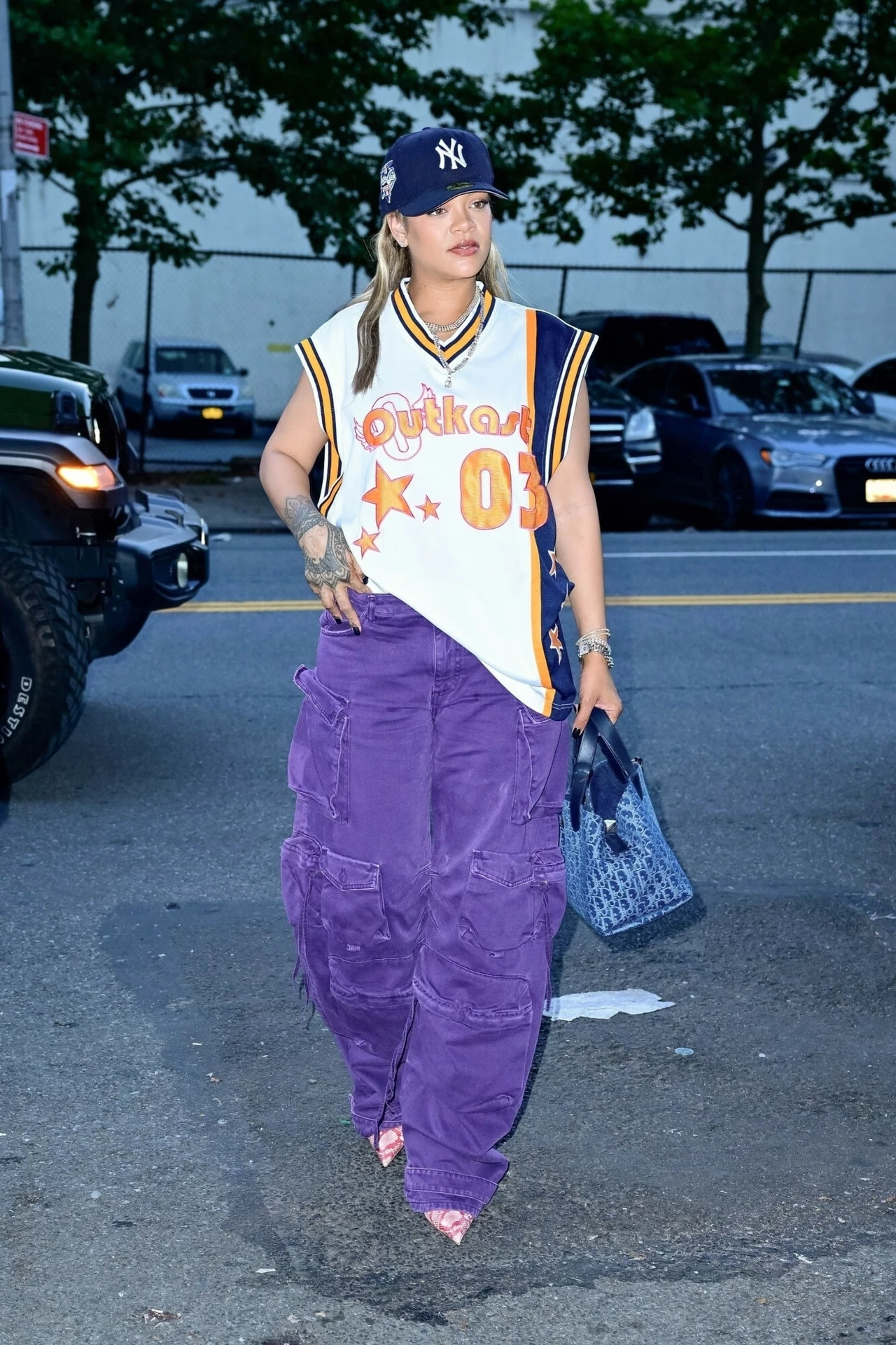 Rihanna w fioletowych spodniach Kobieta w sportowej koszulce bez rękawów z liczbą 03 i napisem Outkast, w szerokich, fioletowych spodniach cargo, z niebieską torebką, czapce z daszkiem New York Yankees i szpilkach, stoi na ulicy na tle zaparkowanych samochodów.