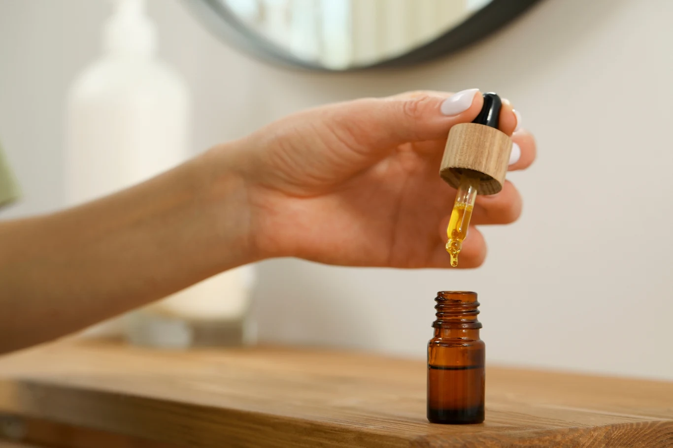 Ręka trzymająca pipetę z kroplą żółtego olejku nad małą buteleczką stojącą na drewnianym blacie, w tle widoczna rozmyta butelka i fragment lustra.