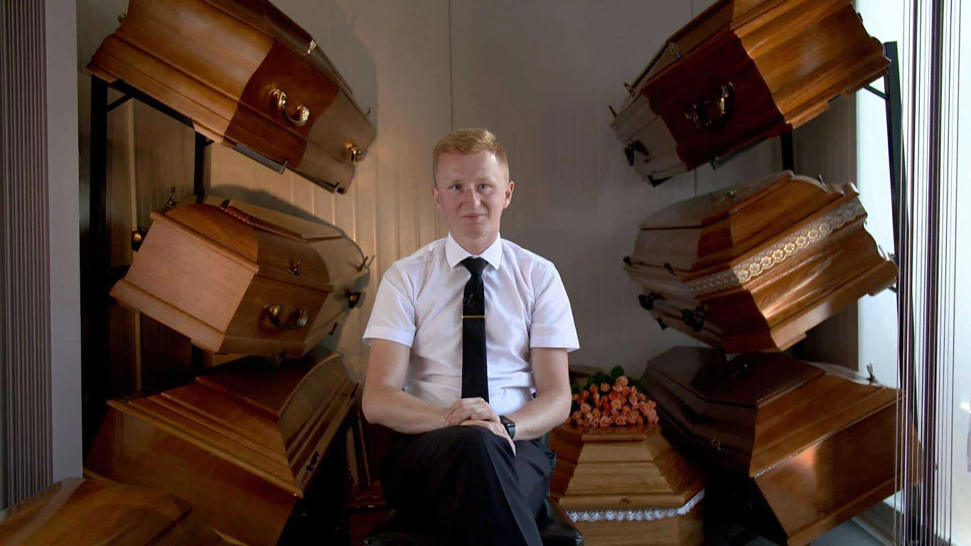 Artur Sierawski pod odejściu ze szkoły otworzył zakład pogrzebowy Młody mężczyzna w białej koszuli i krawacie siedzi na krześle w otoczeniu kilku drewnianych trumien ustawionych na stojakach w pomieszczeniu.