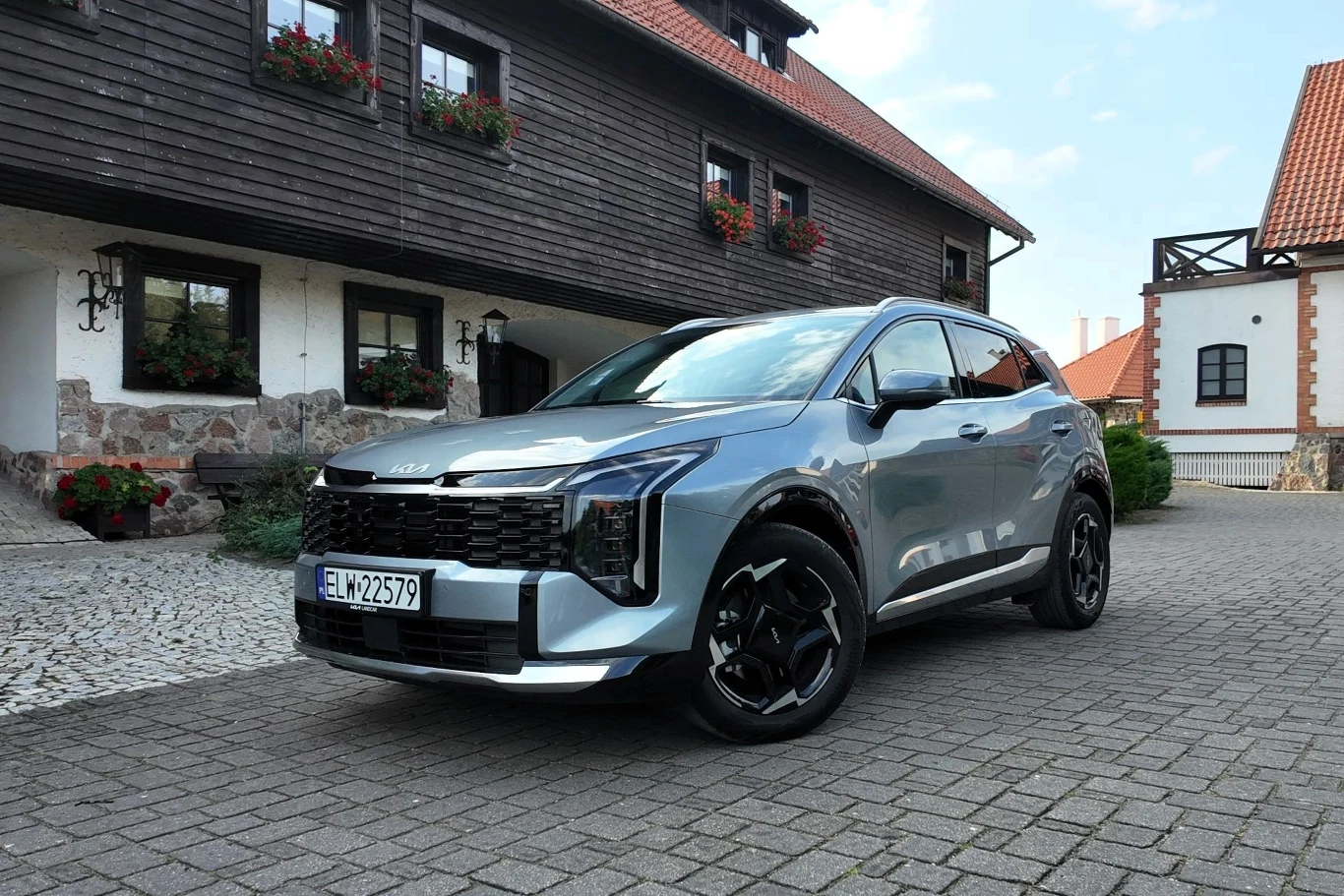 Nowoczesny srebrny SUV zaparkowany na brukowanym dziedzińcu, w tle zabytkowe budynki z drewnianymi i ceglastymi elementami oraz skrzynki z kwiatami pod oknami.