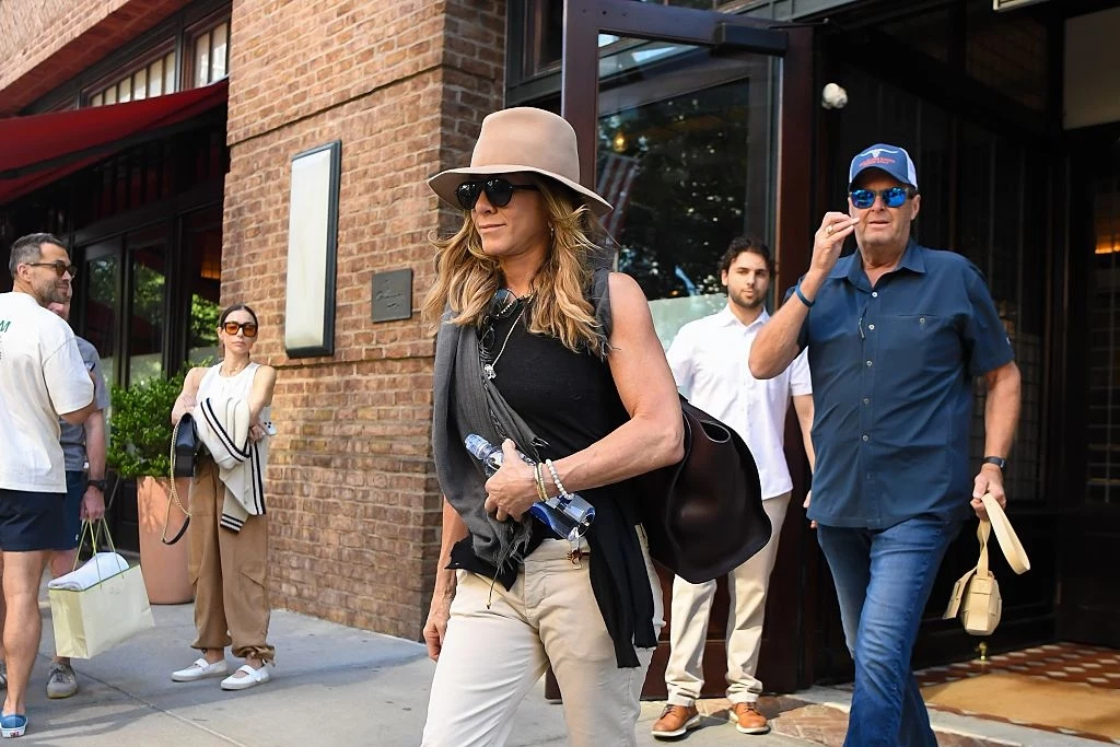 Styl Jennifer Aniston stał się symbolem kalifornijskiej mody Kobieta w kapeluszu i okularach przeciwsłonecznych idzie chodnikiem w mieście, trzymając butelki z wodą i czarną torebkę na ramieniu, za nią kilku ludzi wychodzi z budynku, ubrani w swobodne stroje miejskie.
