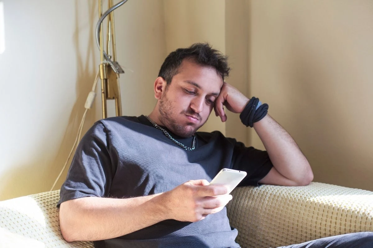 Młody mężczyzna siedzi na fotelu, opierając głowę na ręce, trzymając w drugiej dłoni smartfona, jego wyraz twarzy wskazuje na znudzenie lub znużenie