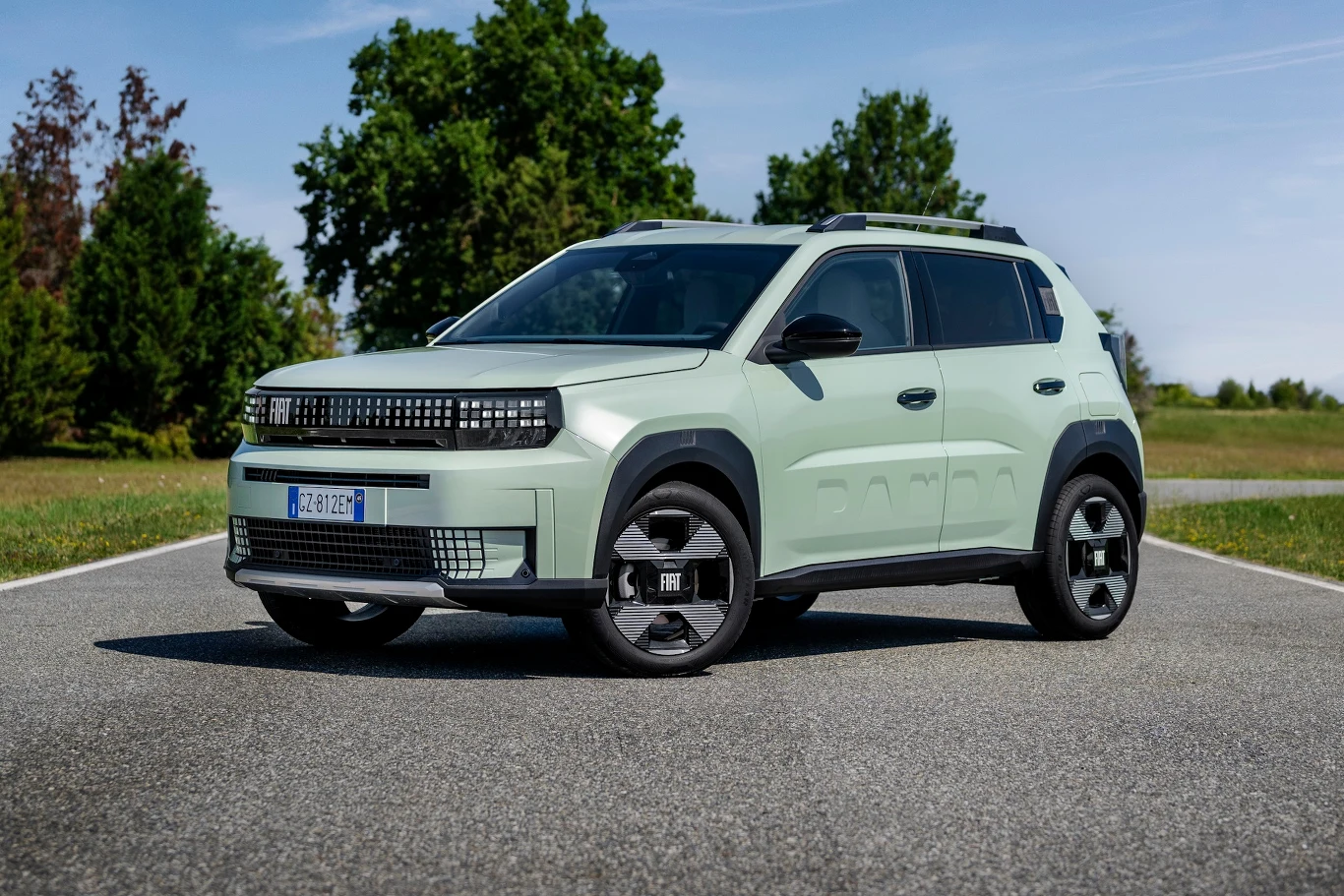 Fiat Grande Panda Nowoczesny, jasny SUV marki Fiat zaparkowany na asfaltowej drodze, otoczony zielenią i drzewami w tle, przy słonecznej pogodzie.