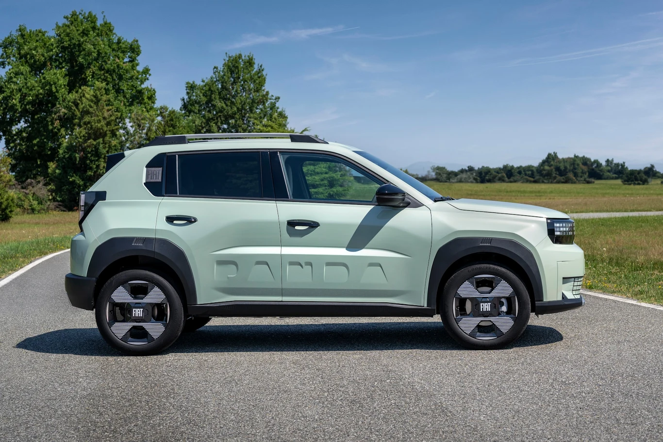 Fiat Grande Panda Jasnozielony nowoczesny samochód osobowy typu SUV stoi na asfaltowej drodze, wokół widoczne są drzewa i trawa, a tło stanowią lekko zamglone wzgórza i niebo pokryte lekkimi chmurami