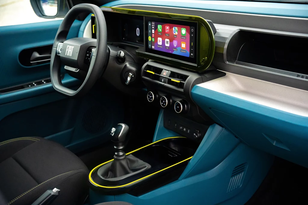Fiat Grande Panda w najbogatszej wersji La Prima Nowoczesne wnętrze samochodu marki FIAT z wyrazistymi turkusowymi i żółtymi akcentami, dotykowym ekranem multimedialnym na desce rozdzielczej, manualną skrzynią biegów oraz ergonomicznymi fotelami.