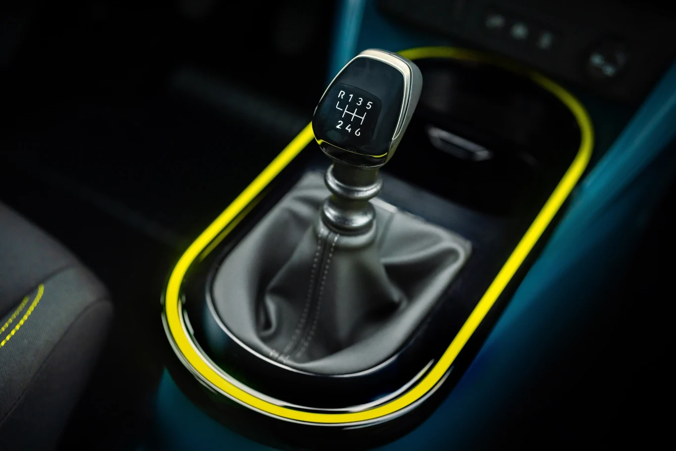 Fiat Grande Panda w wersji spalinowej ma manualną skrzynię biegów Dźwignia zmiany biegów manualnej skrzyni w samochodzie, otoczona żółtym akcentem, z widocznym skórzanym mieszkiem oraz fragmentem fotela kierowcy.