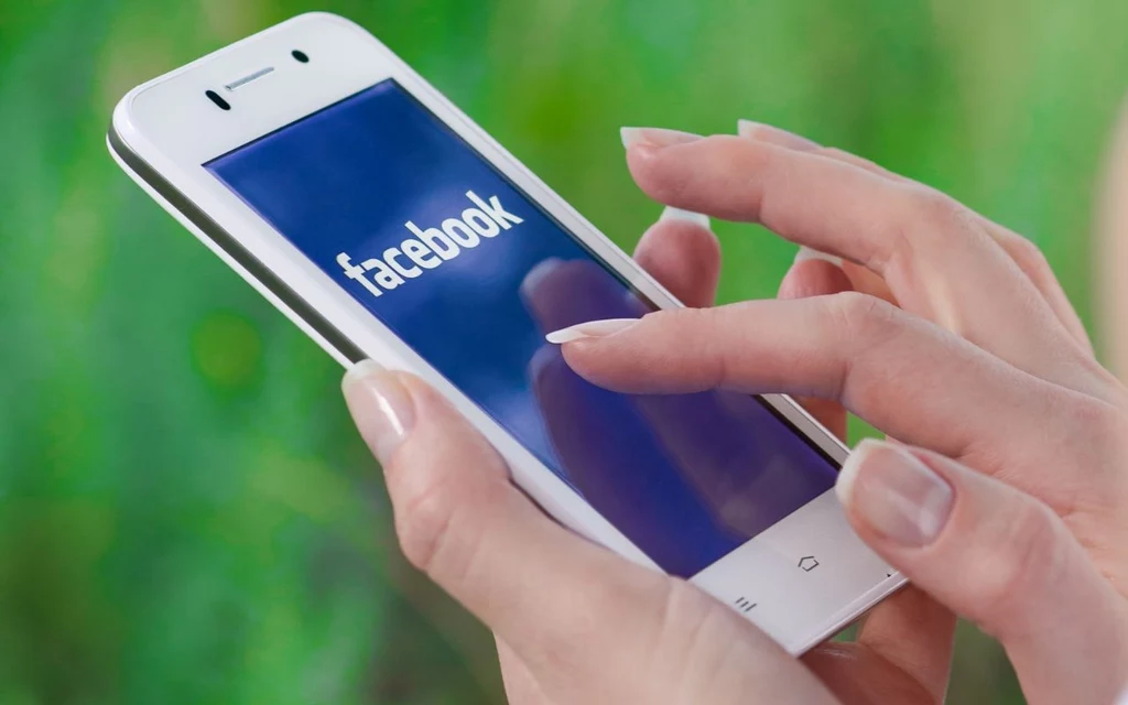 Dłonie kobiety trzymające smartfon z włączoną aplikacją Facebook, wyświetloną na ekranie urządzenia, na tle rozmytego, zielonego otoczenia.