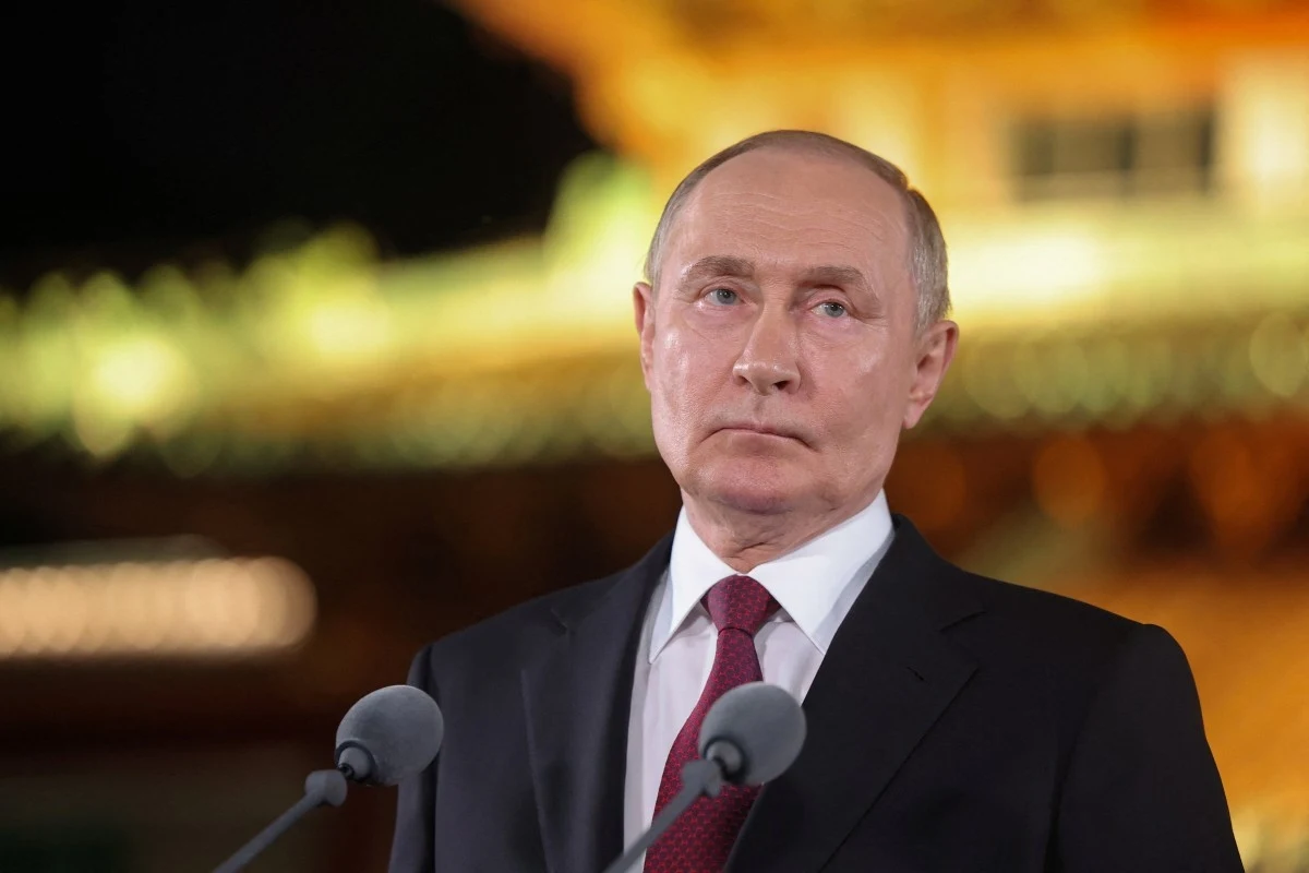 Rosyjski dyktator Władimir Putin w Pekinie Mężczyzna w garniturze i czerwonym krawacie stoi za mównicą z dwoma mikrofonami, w tle rozmyte światła budynku w ciepłych kolorach