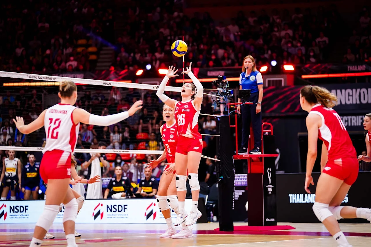 Zawodniczki drużyny siatkówki w czerwonych strojach podczas gry podbijają piłkę przy siatce w hali sportowej, w tle widoczni kibice oraz sędzia stojąca na podwyższeniu.