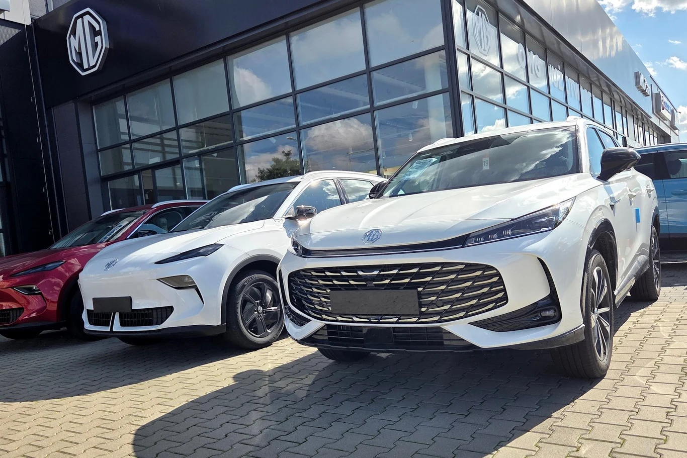 Kilka nowoczesnych samochodów elektrycznych MG ustawionych obok siebie na parkingu przed szklanym salonem samochodowym, wyeksponowane fronty aut podkreślają ich charakterystyczny design i logo marki.